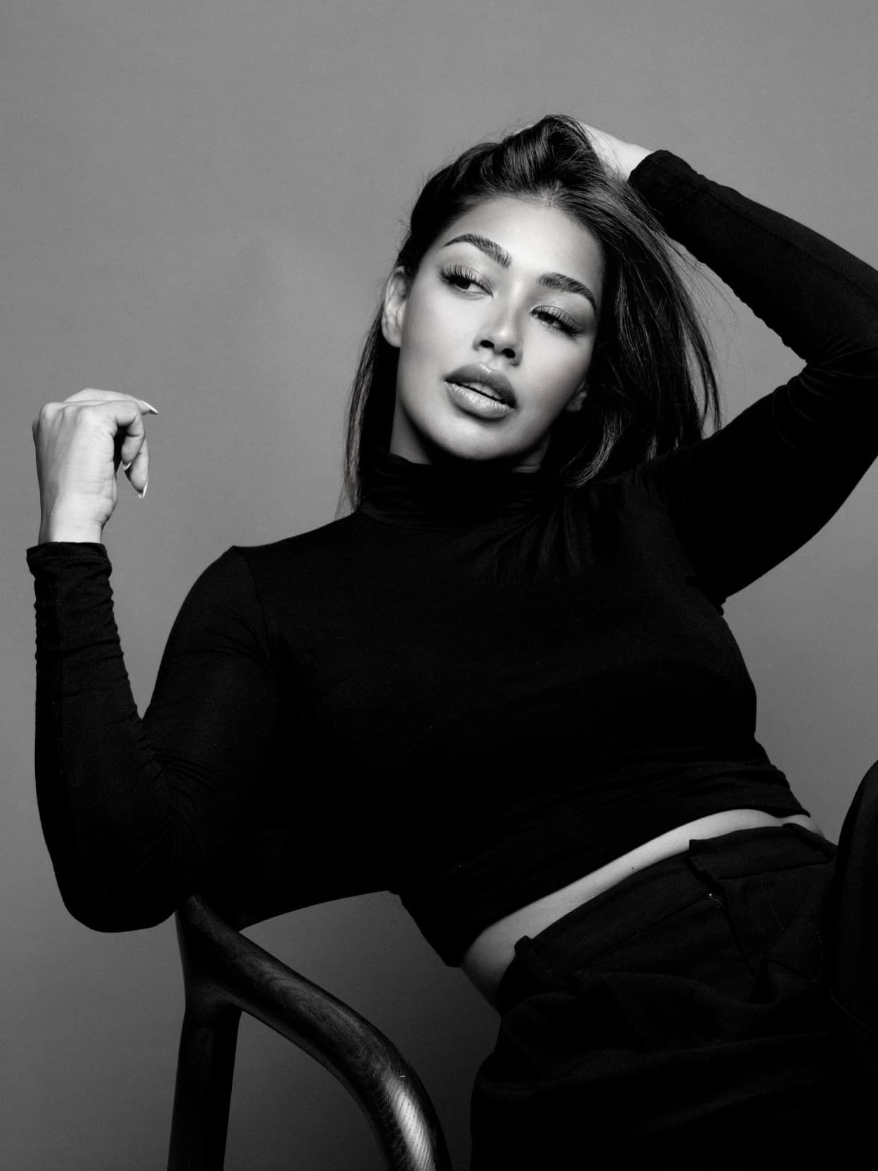 Black and white portrait of Aarti Gayadin seated in a wooden chair, one hand raised in her hair